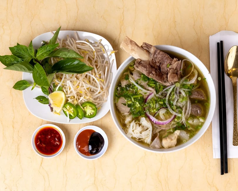 A bowl of pho with a large bone-in beef shank, accompanied by fresh herbs, bean sprouts, and sauces.