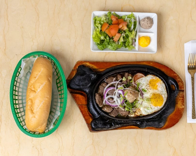 Sizzling beef and eggs with a side salad, pate, mustard, and a baguette in a green basket.