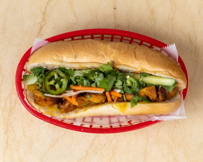 A banh mi sandwich with chicken, cilantro, jalapenos, and pickled vegetables in a red basket.